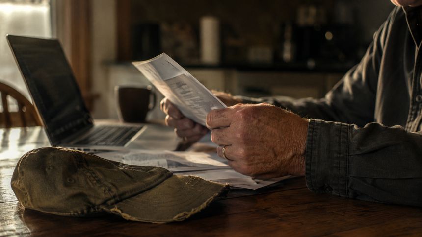 Older man reviewing paperwork at a kitchen table, ball cap beside a laptop