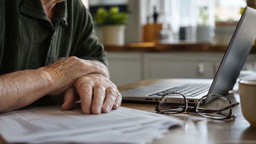 Senior hands resting on printed papers beside reading glasses and a laptop